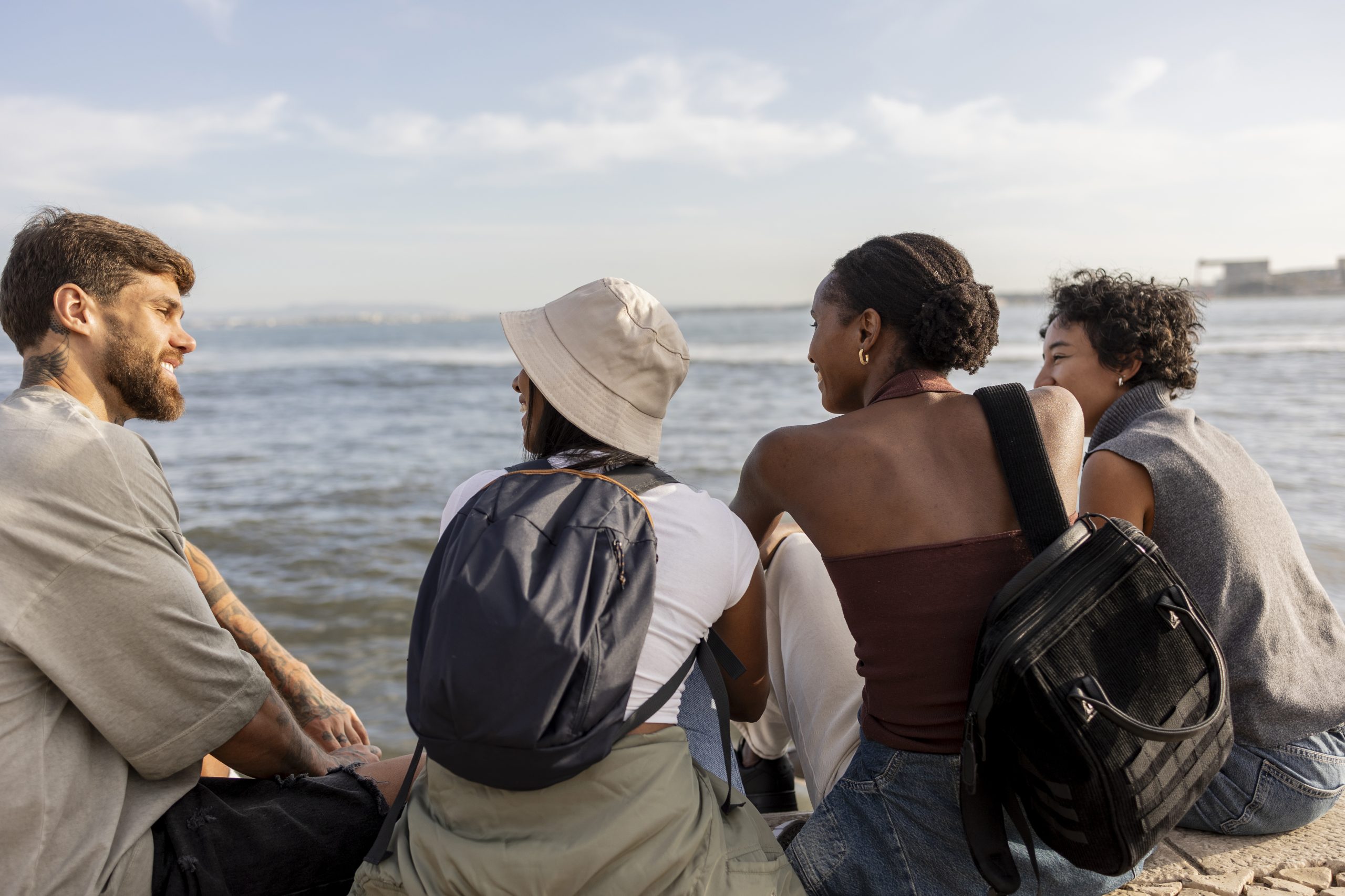 Four young friends, wearing casual clothes and backpacks, are sitting together by the sea, enjoying the view and each other's company on a sunny day